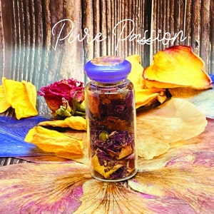 May include: A small glass bottle with a purple lid filled with dried rose petals. The bottle is sitting on a wooden surface with dried rose petals and leaves around it. The text "Pure Passion" is written in white cursive above the bottle.