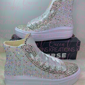 May include: White high-top sneakers covered in iridescent rhinestones and pearls. The sneakers have white laces and a white rubber sole.