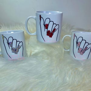 Può includere: Tre tazze in ceramica bianca con un disegno a mano di una mano con unghie lunghe e gioiello. Le unghie sono rosse, rosa e iridescenti. Le tazze sono su una superficie bianca e soffice.