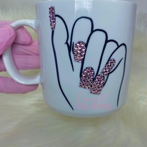 Puede incluir: Taza de cerámica blanca con un contorno negro de una mano con diamantes de imitación rosas en las uñas. La taza tiene el texto "Cereal Kreations" en la parte inferior.