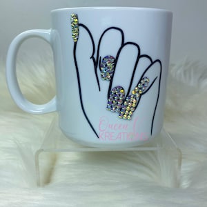 Puede incluir: Taza de cerámica blanca con un contorno negro de una mano con piedras de imitación en las uñas. La taza tiene el texto "Queen L Kreations" en la parte inferior.