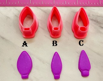 May include: Three pink plastic cookie cutters shaped like light bulbs, labeled A, B, and C. Below each cutter is a purple fondant cutout of a light bulb.