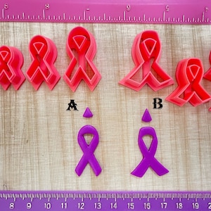 May include: Set of pink and purple plastic ribbon cookie cutters. The set includes five pink cutters in different sizes and two purple cutters. The cutters are arranged on a wooden surface with a ruler.