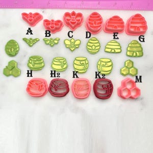 May include: A collection of pink, red, and green bee and honeycomb-shaped cookie cutters. The cutters include various designs such as bees, beehives, and honey pots. The cutters are arranged on a white surface with a pink ruler in the background.