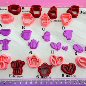 May include: A collection of various coffee-themed cookie cutters in shades of pink, red, and purple. The shapes include coffee cups, a coffee pot, coffee beans, and a teapot. A pink ruler is at the top of the image.