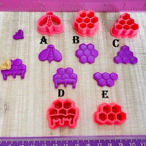 May include: Set of 9 purple and pink plastic cookie cutters shaped like bees and honeycombs. The cutters are arranged on a wooden surface.