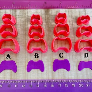 May include: Four sets of pink plastic cookie cutters shaped like a video game controller in four different sizes, labeled A, B, C, and D. Below each set is a purple fondant cutout of a video game controller in the same size as the largest cutter in each set.