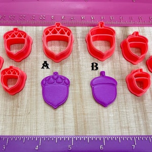 May include: Set of nine red and purple acorn-shaped cookie cutters. The cutters are arranged in two rows on a wooden surface with a ruler in the background. The cutters are different sizes.