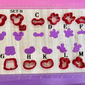 May include: Set of 13 red and purple plastic cookie cutters shaped like Mickey Mouse, Minnie Mouse, and other Mickey Mouse themed shapes. The cutters are arranged on a wooden surface with a ruler in the background. The text "SET-B" is visible in the image.