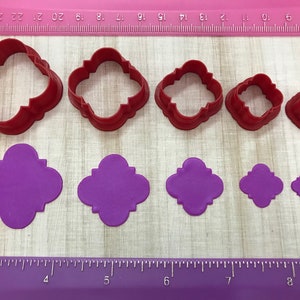 May include: Set of six red plastic cookie cutters in various sizes, shaped like a four-leaf clover with rounded edges. The cutters are shown next to six purple fondant cutouts of the same shape.