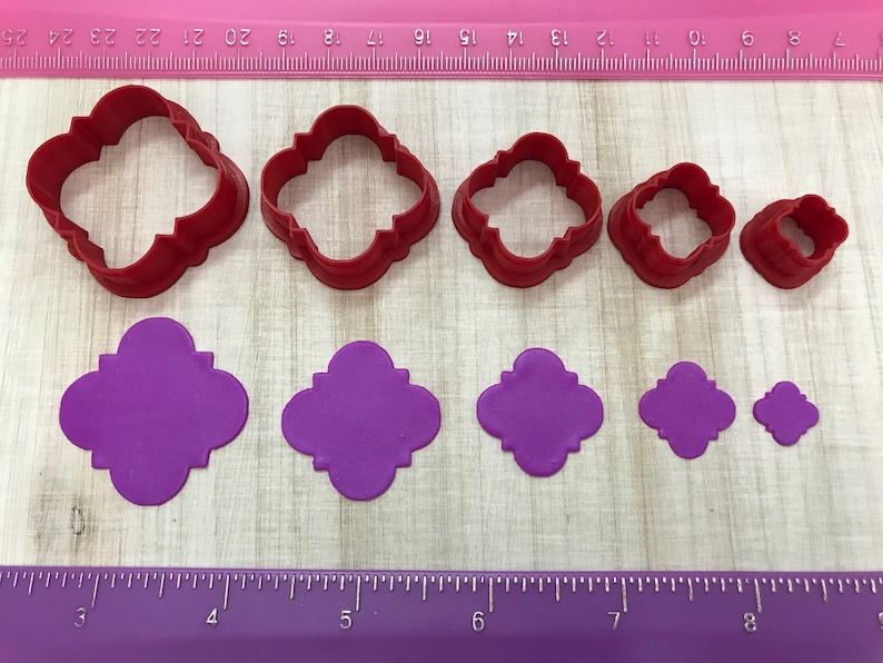 May include: Set of six red plastic cookie cutters in various sizes, shaped like a four-leaf clover with rounded edges. The cutters are shown next to six purple fondant cutouts of the same shape.