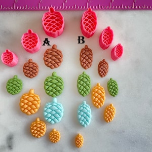May include: A set of 15 pink, green, brown, and blue plastic molds shaped like pine cones. The molds are arranged in a circle on a white surface. The molds are labeled A and B.