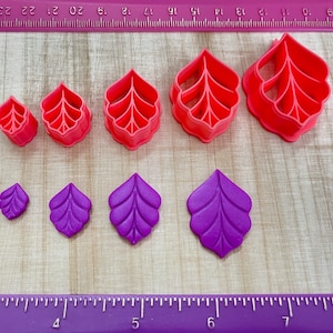 May include: Set of seven pink and purple leaf-shaped cookie cutters. The cutters are arranged in two rows, with the larger cutters in the top row and the smaller cutters in the bottom row. The cutters are made of plastic and have a smooth finish.