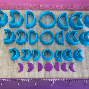 May include: A set of 18 blue plastic cookie cutters in various crescent moon and circle shapes. The cutters are arranged in three rows on a wooden surface. There are four purple fondant crescent moons in the bottom row.