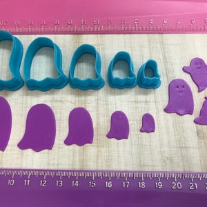 May include: A set of 10 purple and blue plastic cookie cutters in the shape of ghosts, pumpkins, and witches' hats. The cutters are arranged on a wooden surface with a ruler for scale.