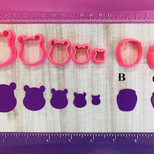 May include: Five pink plastic cookie cutters in the shape of a hippopotamus, with the largest cutter measuring approximately 2 inches in diameter. Three purple fondant cutouts of a hippopotamus, with the largest cutout measuring approximately 2 inches in diameter. Two pink plastic cookie cutters in the shape of a jar, with the largest cutter measuring approximately 2 inches in diameter. Two purple fondant cutouts of a jar, with the largest cutout measuring approximately 2 inches in diameter.