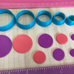 May include: A set of seven blue plastic cookie cutters and eight circles of red and purple fondant. The cookie cutters are in various sizes and shapes, including circles, squares, and rectangles. The fondant circles are also in various sizes.