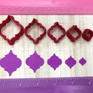 May include: Set of six red plastic cookie cutters in various sizes, shaped like a quatrefoil, and six purple paper cutouts of the same shape.