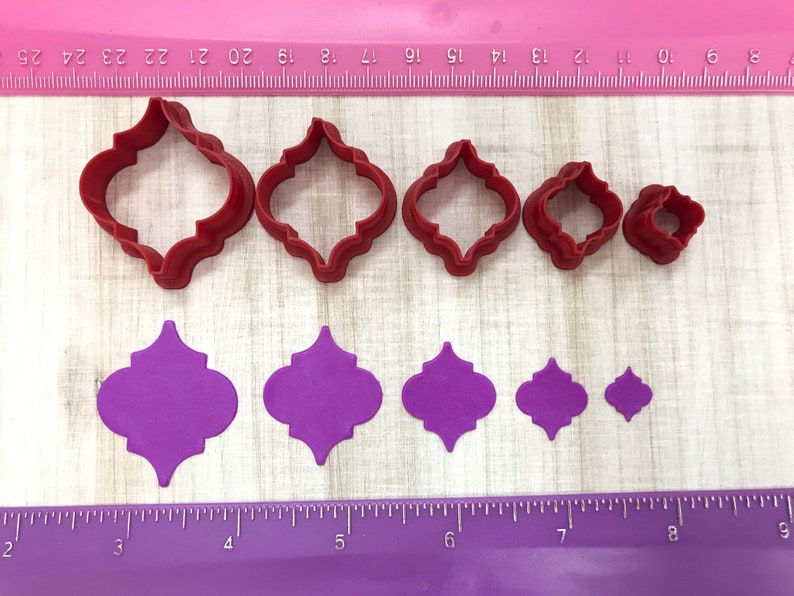 May include: Set of six red plastic cookie cutters in various sizes, shaped like a quatrefoil, and six purple paper cutouts of the same shape.