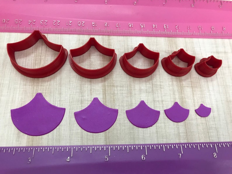 May include: Set of seven red and purple plastic cookie cutters shaped like a fan. The cutters are arranged in order of size from largest to smallest. A ruler is visible in the background.