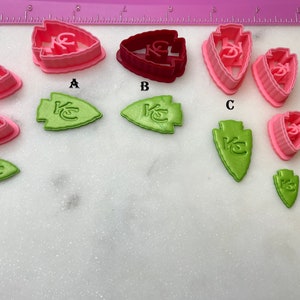 May include: A set of nine cookie cutters in various sizes. The cutters are shaped like an arrowhead with the letters "KC" inside. The cutters are in three colors: pink, red, and green.