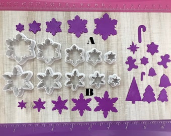 May include: A set of 12 purple and white plastic cookie cutters in various shapes, including snowflakes, a candy cane, a bell, a star, a Christmas tree, and a gingerbread man. The cutters are arranged on a wooden surface with a ruler in the background.
