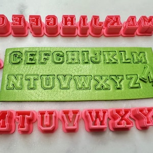 May include: A set of pink plastic alphabet cookie cutters and a green plastic alphabet embossing mat. The cutters are in uppercase letters and the mat has both uppercase and lowercase letters.