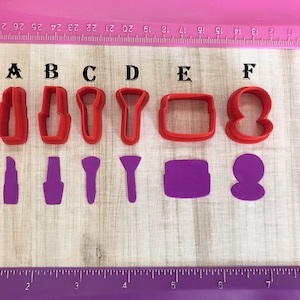 May include: Six red plastic cookie cutters shaped like lipstick, a makeup brush, a compact, and a powder puff. Below the cutters are six purple plastic cookie cutters shaped like the same items.