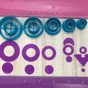 May include: A set of 10 cookie cutters in two colors: teal and purple. The teal cutters are in a variety of circle shapes, some with a cross-hatch design. The purple cutters are in a variety of circle and geometric shapes.