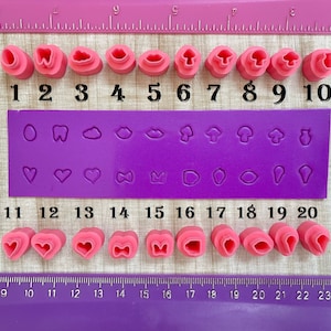 May include: A set of 20 pink polymer clay stamps with various designs, including hearts, mushrooms, clouds, lips, and teeth. The stamps are numbered 1 through 20 on a purple background.