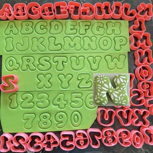 May include: A collection of pink plastic cookie cutters in the shapes of letters and numbers, arranged alongside two large light green silicone mats embossed with the full alphabet and digits zero through nine, plus one green letter N cookie.
