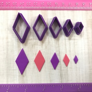 May include: A collection of purple diamond-shaped cookie cutters in various sizes, alongside diamond-shaped cutouts in purple and pink. The items are arranged on a wooden surface with a pink ruler in the background.