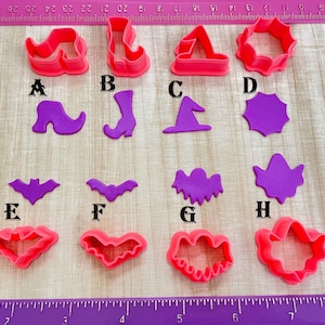 May include: A set of ten pink and purple plastic cookie cutters shaped like Halloween characters, including a witch's hat, a bat, a ghost, and a boot. The cutters are arranged on a wooden surface with a ruler.
