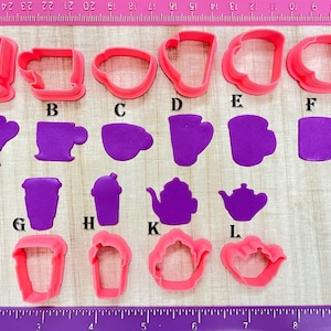 May include: A set of 13 pink and purple plastic cookie cutters shaped like different types of coffee cups, teacups, and teapots. The cutters are arranged on a wooden surface with a ruler in the background.