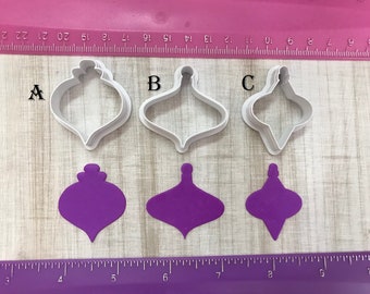 May include: Three white plastic Christmas ornament cookie cutters, labelled A, B, and C, with three purple fondant ornaments cut out using the cutters.