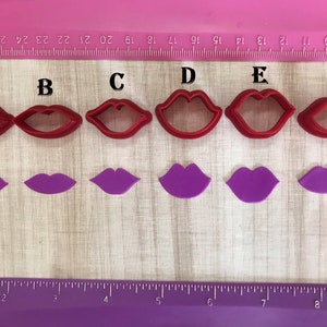 May include: A collection of six red plastic lip-shaped cookie cutters, each with a different design. Below each cutter is a matching purple lip-shaped cutout. The items are arranged on a pink surface with a ruler along the top edge.