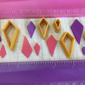 May include: An assortment of diamond-shaped craft supplies, including purple and pink cutouts and yellow plastic cutters. The items are arranged on a wooden surface with a purple ruler, ideal for crafting and DIY projects.