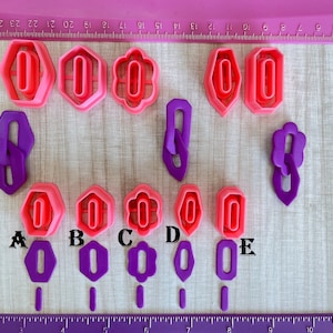 May include: A set of nine pink and purple plastic cookie cutters in various shapes, including circles, ovals, and flowers. The cutters are arranged on a pink and white surface with a ruler in the background.