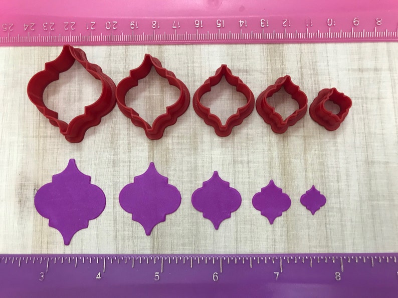 May include: Set of six red plastic cookie cutters in various sizes, shaped like a quatrefoil, with a purple fondant cutout of the same shape in the background.