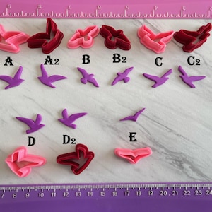 May include: Assortment of pink and burgundy bird-shaped cookie cutters and purple bird-shaped fondant decorations. The cutters are labeled A, A2, B, B2, C, C2, and the fondant birds are labeled D, D2, and E. A pink ruler is in the background.