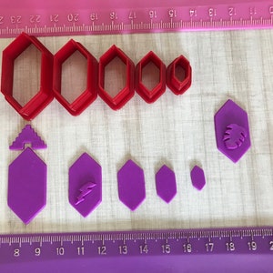 May include: A collection of red and purple polymer clay cutters in various sizes and shapes. The red cutters are geometric, while the purple cutters have decorative shapes, including a lightning bolt and a leaf. The cutters are arranged on a pink surface with ruler markings.