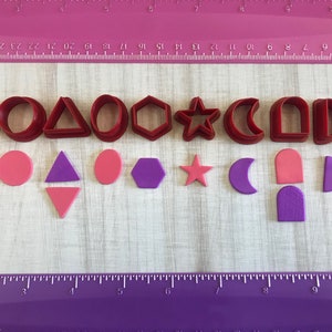 May include: A set of 10 red plastic cookie cutters in various shapes, including a square, circle, triangle, oval, pentagon, star, crescent moon, arch, half circle, and a rectangle. The cutters are arranged in two rows on a white surface with a purple ruler in the bottom of the image.