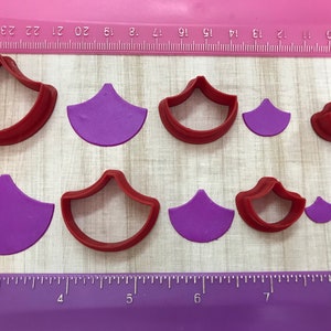 May include: Set of nine red and purple plastic cookie cutters shaped like a scallop shell. The cutters are arranged in two rows on a wooden surface with a ruler in the background.