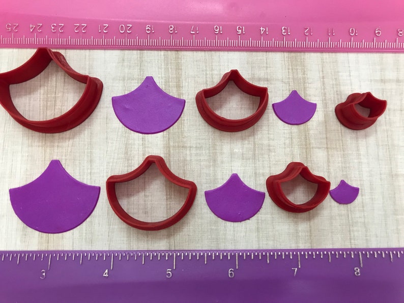 May include: Set of nine red and purple plastic cookie cutters shaped like a scallop shell. The cutters are arranged in two rows on a wooden surface with a ruler in the background.