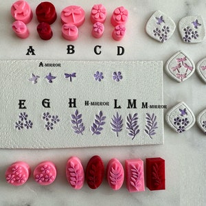 May include: A set of 12 polymer clay stamps featuring various floral and dragonfly designs. The stamps are arranged in rows labeled A-M, with some stamps mirrored. The stamps are pink, red, and purple.