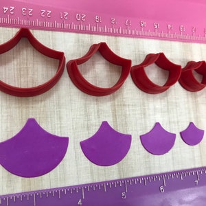 May include: Set of six red and purple plastic cookie cutters shaped like a scallop shell. The cutters are arranged in order of size, with the largest cutter at the top and the smallest cutter at the bottom. The cutters are on a pink and white ruler.