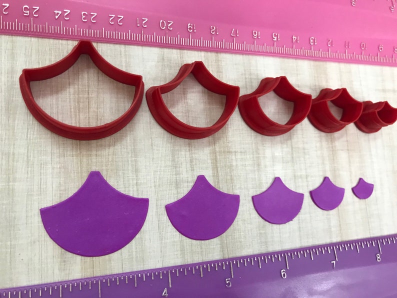 May include: Set of six red and purple plastic cookie cutters shaped like a scallop shell. The cutters are arranged in order of size, with the largest cutter at the top and the smallest cutter at the bottom. The cutters are on a pink and white ruler.