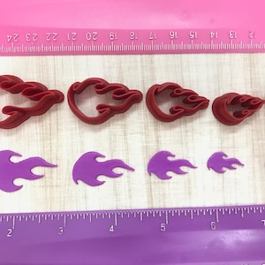 May include: A set of eight red and purple plastic cookie cutters shaped like flames. The cutters are arranged in two rows on a pink and purple surface with a ruler for size reference.