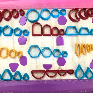 May include: A set of colorful plastic cookie cutters in various shapes, including circles, ovals, hexagons, triangles, and crescent moons. The cutters are arranged on a wooden surface with a ruler.