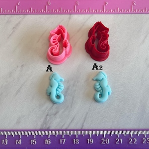 May include: An overhead shot displays four seahorse-themed items. Two are light blue seahorse figurines, and the other two are seahorse-shaped cookie cutters, one pink and one red. The items are arranged on a light-colored surface with pink and purple rulers.